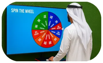 Arab male playing Spin the Wheel game at an exhibition event in Dubai, wearing traditional kandura, fully branded setup with digital screen and prize display, engaging crowd.